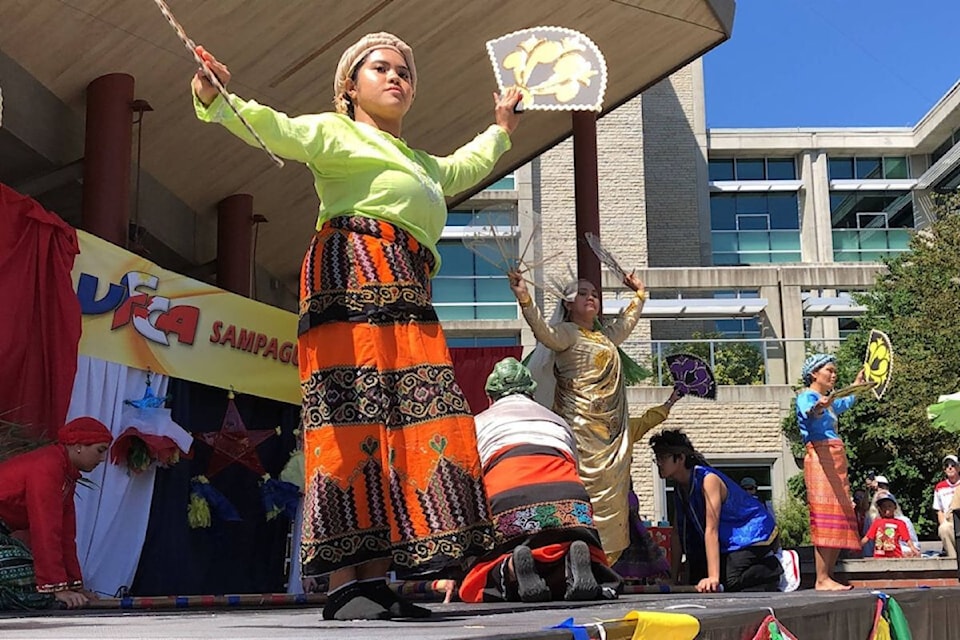 Young woman in traditional lime green top and patterned orange and black skirt performing cultural dance outdoors, holding circular prop and stick with arms outstretched, with other performers and festival banners visible in background.